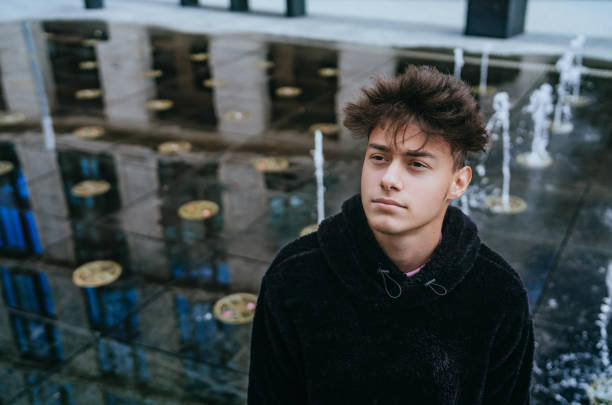 istock young man in a black hoodie stands thoughtfully by a reflective water feature with small fountains, creating an artistic and moody urban scene 2153832900