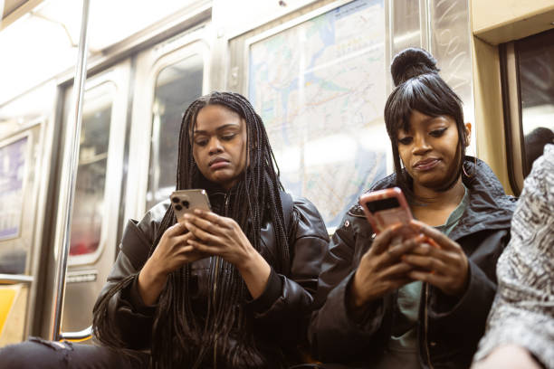 Two young women, reflective of a diverse urban population, navigate their smartphones while seated in a New York City subway car, dressed in casual attire suitable for travel.