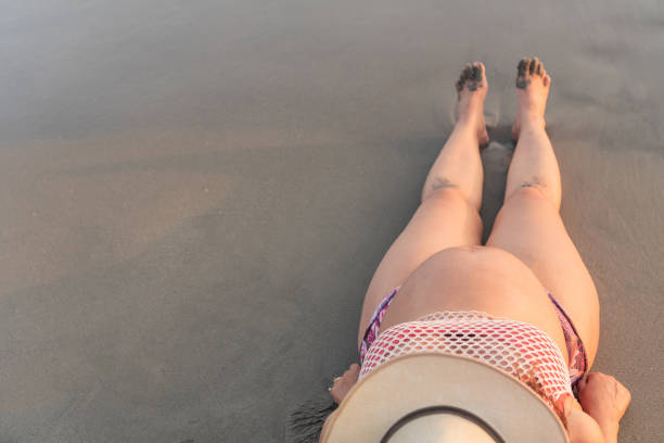 Top view of an expectant mother lying on the sand, showcasing her baby bump, wearing a hat and relaxing by the shore.