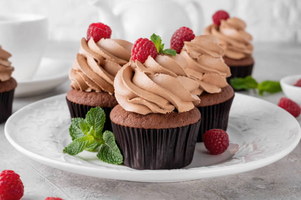 istock Chocolate cupcakes with cream decorated with fresh raspberries and mint leaves on a gray concrete background. Delicious holiday dessert 2153866743
