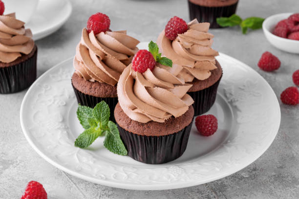 istock Chocolate cupcakes with cream decorated with fresh raspberries and mint leaves on a gray concrete background. Delicious holiday dessert 2153866746