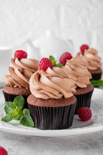 istock Chocolate cupcakes with cream decorated with fresh raspberries and mint leaves on a gray concrete background. Delicious holiday dessert 2153866759