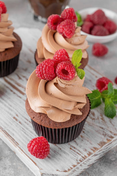 istock Chocolate cupcakes with cream decorated with fresh raspberries and mint leaves on a gray concrete background. Delicious holiday dessert 2153866766
