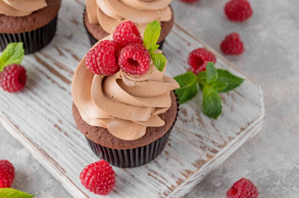 istock Chocolate cupcakes with cream decorated with fresh raspberries and mint leaves on a gray concrete background. Delicious holiday dessert 2153866767
