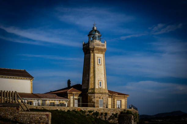 A photo of the San Juan de Aviles lighthouse, a towering white structure with a red lantern room, standing proudly on a rocky outcrop overlooking the vast expanse of the salt flats. The sun casts a warm glow over the scene, illuminating the intricate details of the lighthouse's architecture and the shimmering surface of the salt flats. In the distance, the hazy outline of mountains adds a touch of grandeur to the panorama.