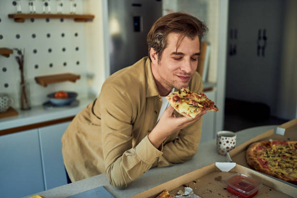 istock A smiling man leaning on a kitchen counter and enjoying a slice of pizza 2153935297