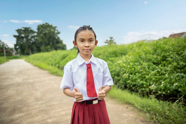 Young female Indonesian with elementary school uniform going to school by walking and giving thumbs up against green scenery as background. Back to school concept.