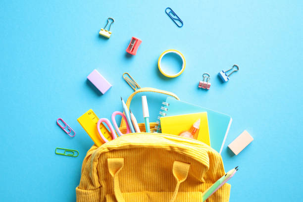 istock Yellow backpack with an array of colorful school supplies spilling out, including notebooks, pencils, and scissors on blue background. Back to school concept. 2153958497