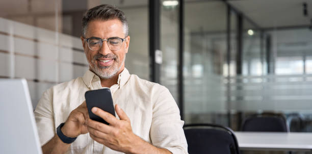 istock Banner with middle-age Hispanic business manager ceo using cell phone mobile app. Smiling Latin or Indian mature man businessman holding smartphone in office, working as trader on pc, with copy space 2153961610