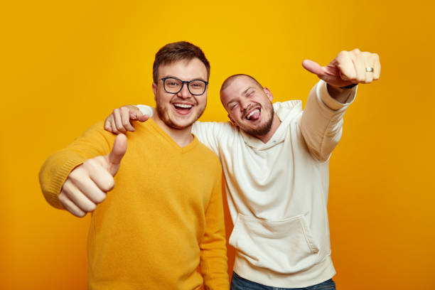 istock Image of two cheerful handsome guys embracing each other while showing thumbs up against yellow wall 2153967939