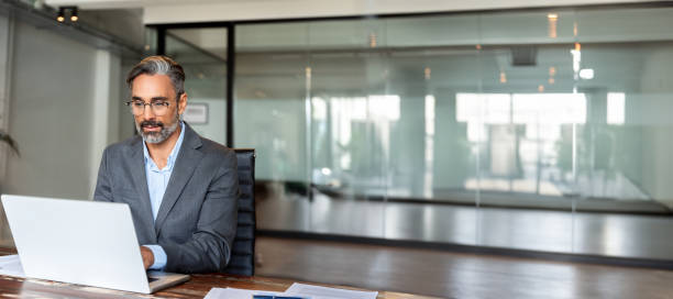 istock Mature Indian or Latin business man ceo trader using computer, typing, working in modern office, doing online data market analysis, thinking planning tech strategy looking laptop. Banner, copy space 2153986415