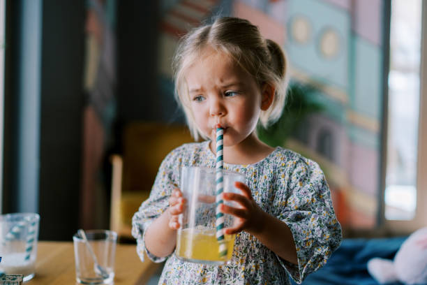 istock Little girl drinks juice from a glass through a straw while standing at a table in a cafe 2153988450