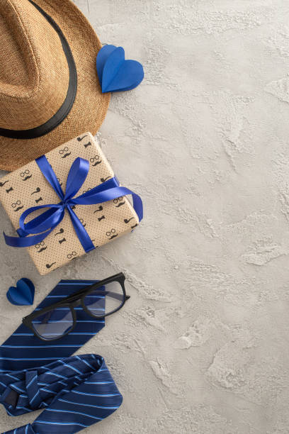 istock A stylish Father's Day arrangement featuring a straw hat, blue necktie, gift box with blue ribbon, glasses, and paper hearts on a vertical textured concrete background 2153989055