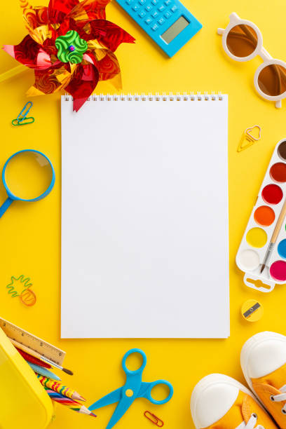 istock Creative summer program theme. Top vertical  view of art materials: sketchpad, colored pencils, paints, clips, calculator, magnifying glass, sunglasses, sneakers, pinwheel on yellow background with copy space 2153989105