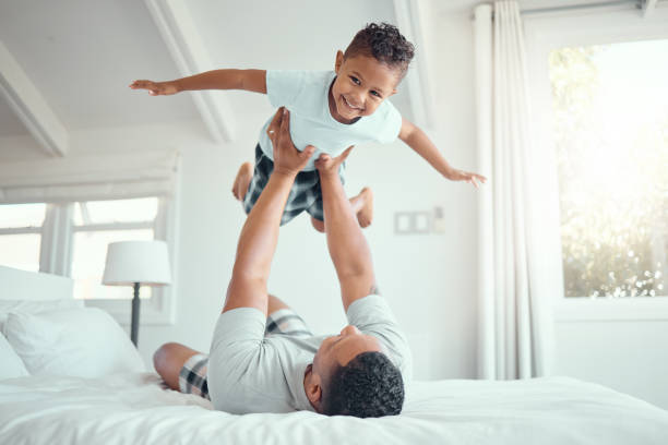 istock Cute little mixed race boy lying in fathers arms while looking at the camera and smiling. Loving family with father lifting happy little child son playing plane and having fun on bed 2153989601