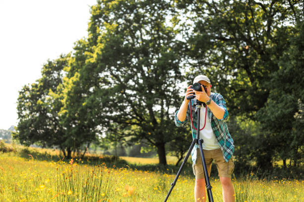 Portrait of a mid adult male photographer at work in the field, surrounded by beautiful nature. The man has his DSLR camera mounted on a tripod. The man is wearing casual clothing - khaki shorts, a green check shirt over a white t shirt, and a beige baseball cap on his head. The man has a contented expression on his face as he works on creating images and video.
