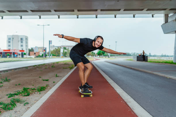 A happy male skateboarder enjoying a ride with his arms outstretched under an overpass, wearing black sports attire and headphones