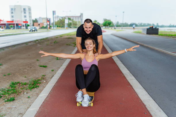 A happy young couple is engaging in a playful activity where the man pushes the woman on a skateboard in an open area
