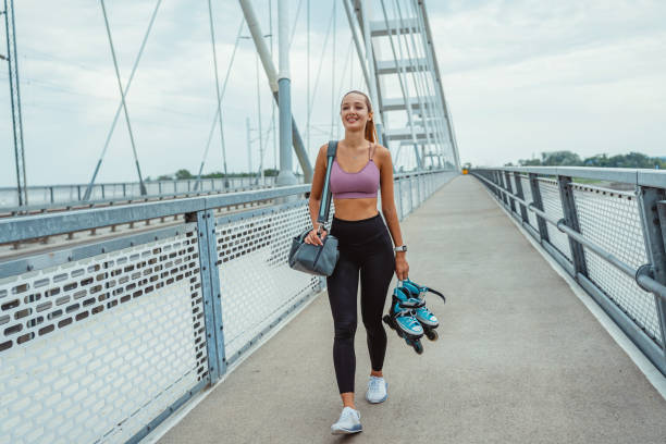 A happy, fit woman in workout attire walks on a modern pedestrian bridge, carrying a gym bag and jacket