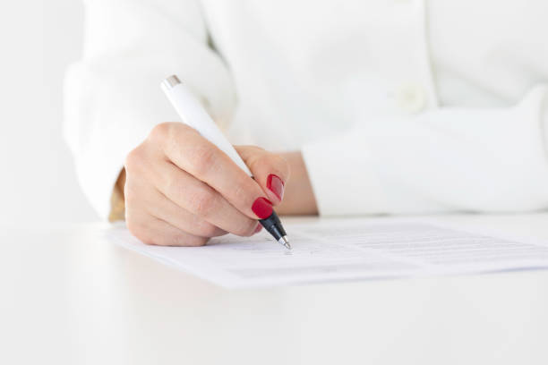 Unrecognizable caucasian woman wearing white jacket and red nail polish is signing contract paper.