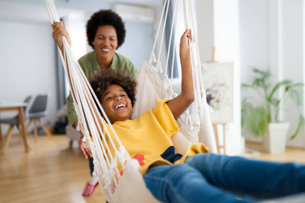 Affectionate mother swinging her female child on a swing Cheerful African American young girl playing on a rope swing in the living room at home. Affectionate mother swinging her female child on a swing. swinger photo stock pictures, royalty-free photos & images