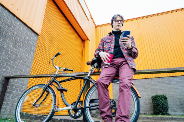 istock Young hipster man using mobile phone while sitting on cruiser bicycle near orange wall. Student using 5g network,rests after riding. Eco friendly transport for carbon footprint. Sustainable lifestyle 2154044259