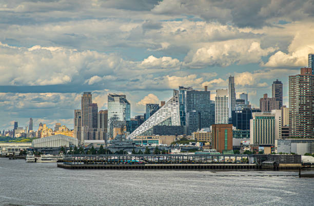 New York, NY, USA - August 1, 2023: Triangle shaped white apartment complex on west 57th. Intrepid plane carrying vessel museum on pier 67 towers over rest. Blue cloudscape and urban jungle of high-rise buildings
