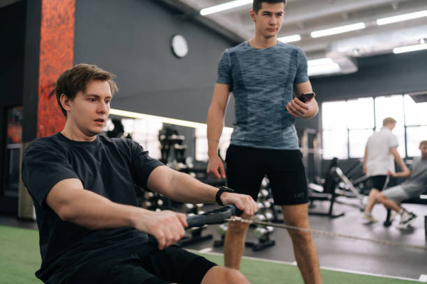 Focused beginner sportsman with personal instructor working out in gym by using rowing machine. Sweating man doing exercises for back muscles under supervision of trainer