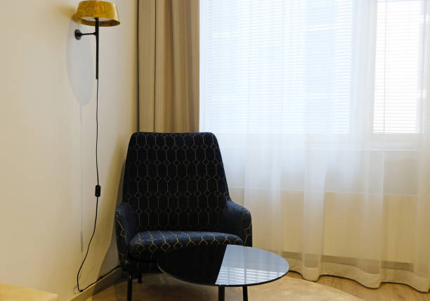 Modern interior design. Black armchair, black glass table, wall lamp, blinds on windows with white curtains. Stylish comfortable furniture. Minimalism.