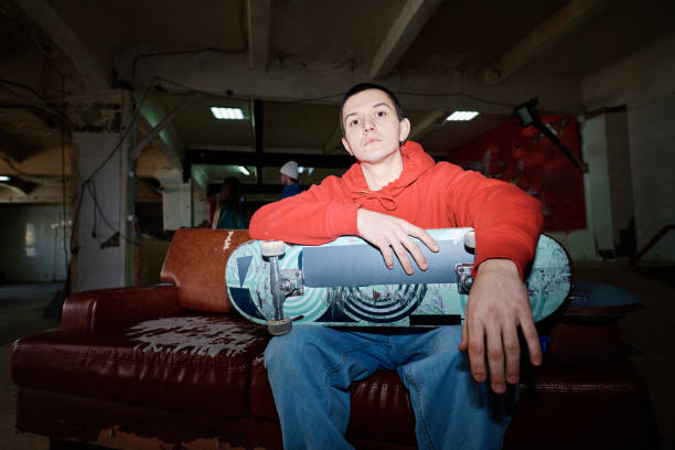 Flash photo of self-confident gen Z man wearing red hoodie and jeans sitting on shabby couch in skatepark looking at camera