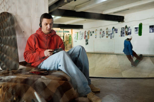Medium long shot of gen Z guy wearing headphones relaxing in skatepark turning on old portable player, copy space