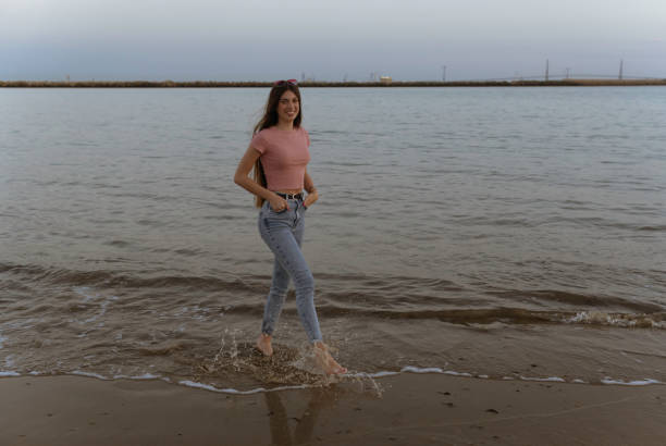 A woman is walking in the water wearing a pink shirt and blue jeans. The water is calm and the sky is cloudy