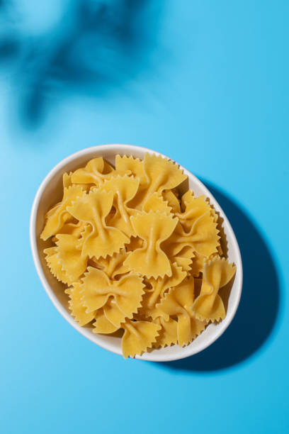 istock Uncooked bow tie pasta, farfalle in a white bowl on a blue background with shadows 2154124194