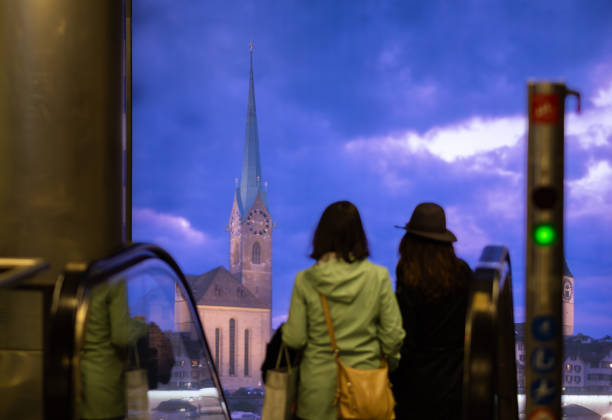 Zurich, Switzerland - May 16, 2024: Two individuals on escalator, against urban view of Zurich with historic church and dramatic sky.