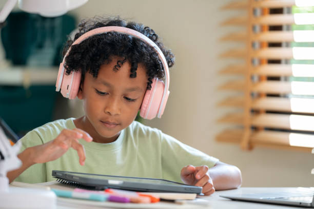 African elementary student wearing headphones appears bored and disengaged while studying e-learning online at home in the living room. Despite the modern technology and access to education, he lacks happiness and enthusiasm for his studies