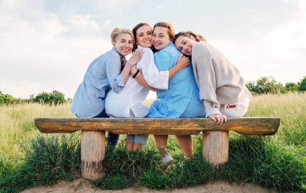Four smiling women dressed light summer clothes harm embracing sitting on meadow bench during outdoor walking and looking at camera. Woman's friendship, relations, and happiness concept image.