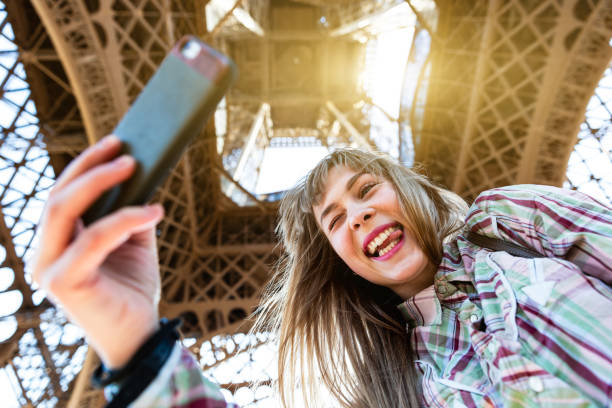 Beautiful blonde girl taking a selfie under the Eiffel Tower in Paris - Young woman smiling and grimacing with the tongue out of the mouth - Travel and lifestyle concepts