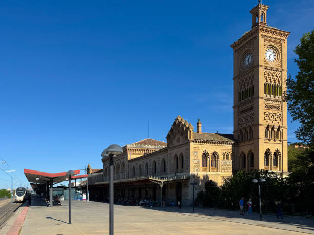 Wide shot of the Toledo train station. Long view with the clock tower in the foreground and the mudejar-style station just behind. High-speed Renfe train the background. Late afternoon, clear blue sky, strong sun.