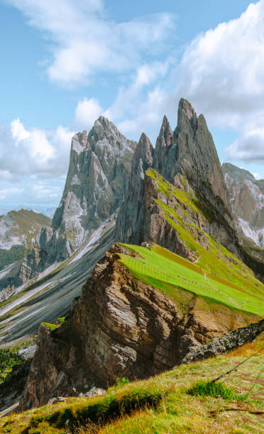 tunning springtime view of Seceda in the Dolomites, Italy. Under a sunny day with a picturesque cloudscape, the green meadows burst into bloom, creating a vibrant landscape. The majestic mountain slopes bask in sunlight, showcasing the area's natural beauty. Known as a popular ski center, Seceda transforms into a verdant paradise during spring, offering a captivating scene for nature lovers and adventure seekers alike.