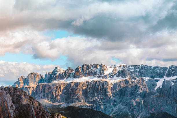 A breathtaking view of the snow-covered mountain range in the Dolomites, Italy, illuminated by the warm glow of sunset. The scene is enhanced by clouds draped over the peaks, adding a mystical and serene atmosphere to the rugged landscape. This stunning natural beauty captures the essence of the Italian Alps, making it perfect for themes related to nature, travel, outdoor adventure, and the enchanting allure of mountain vistas