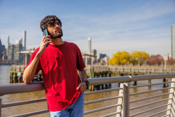 Phone conversation by the river. Pakistani man remote working at sunny pier on East River in Queens, Long Island City