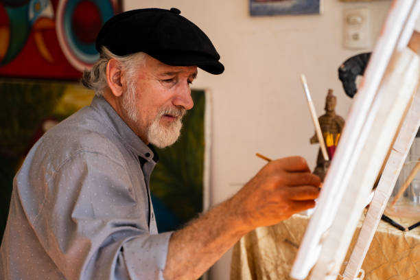Man between 50-60 with gray hair and a large white beard, dressed in a French beret and comfortable and cool clothes, facing the camera in profile while making art and painting a beautiful painting in his studio, mixing paints, creating tones and giving brush strokes to finish your work