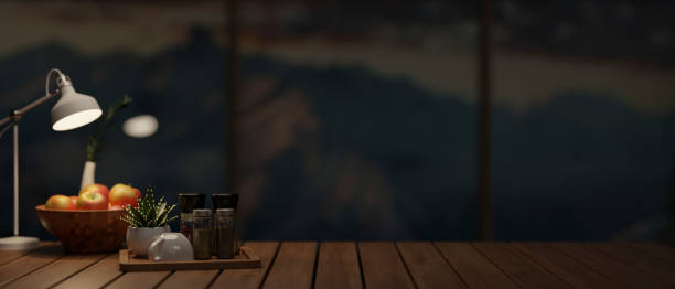 A wooden tabletop featuring a coffee cup, spice bottles, a potted plant, an apple bowl, a table lamp, and a space for displaying products in a dark room at night. 3d render, 3d illustration