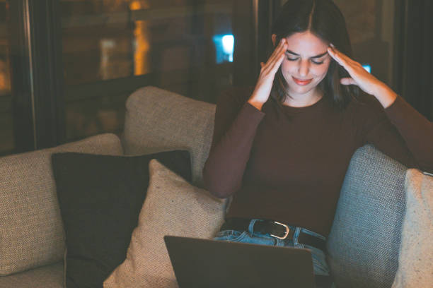 istock One woman massaging her heads for strong migraine headache sitting on the sofa after using laptop by night in online activity. People suffering from head pain and stress for failure. Head ache disease 2154243636