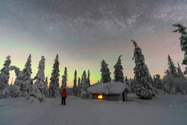 Tourist standing in the frozen forest by the mountain hut  covered with snow under northern lights, Finland, Lapland, Scandinavia, Europe
