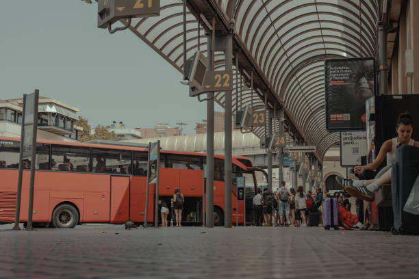 The image shows a bus station featuring several bus bays, each marked by numbered signs hanging from the ceiling. Red buses are parked at some of the bays, and a group of people can be seen either waiting or walking around the station. The station has a curved metal roof that provides shade for the area. On the right side of the image, there is a digital advertisement board displaying an ad with text and an image of a person’s face.