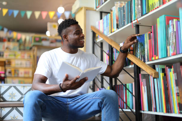 istock Young African student studying in a university library, holding a book, sitting by a bookshelf. 2154280466