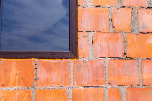 Detailed close-up of an unfinished brick wall with a window reflecting the cloudy sky, emphasizing construction and urban development.