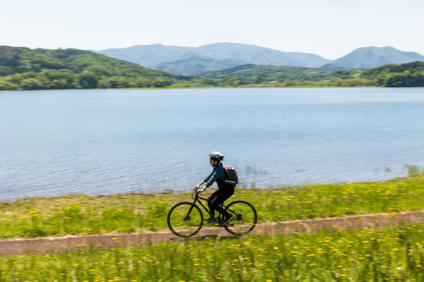 A young Japanese woman riding her bicycle and wearing a helmet through the countryside by a large lake.