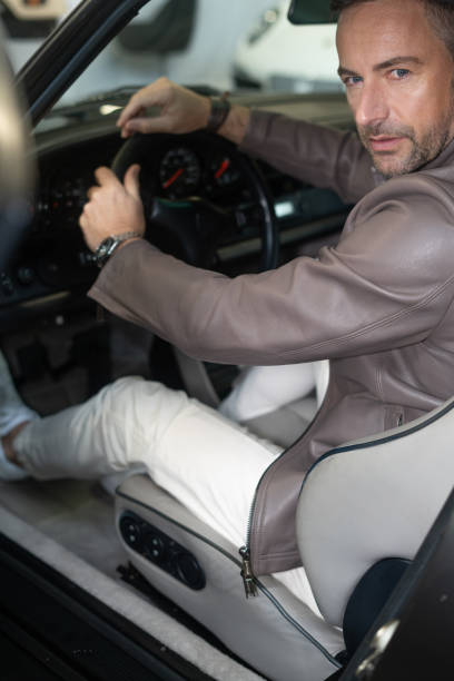 Sportlicher Mann mittleren Alters mit grauen Schläfen im hochwertigem Outfit sitzt in einem Sportwagen mit Ledersitzen und schaut in die Kamera, beige, weiß, Desig